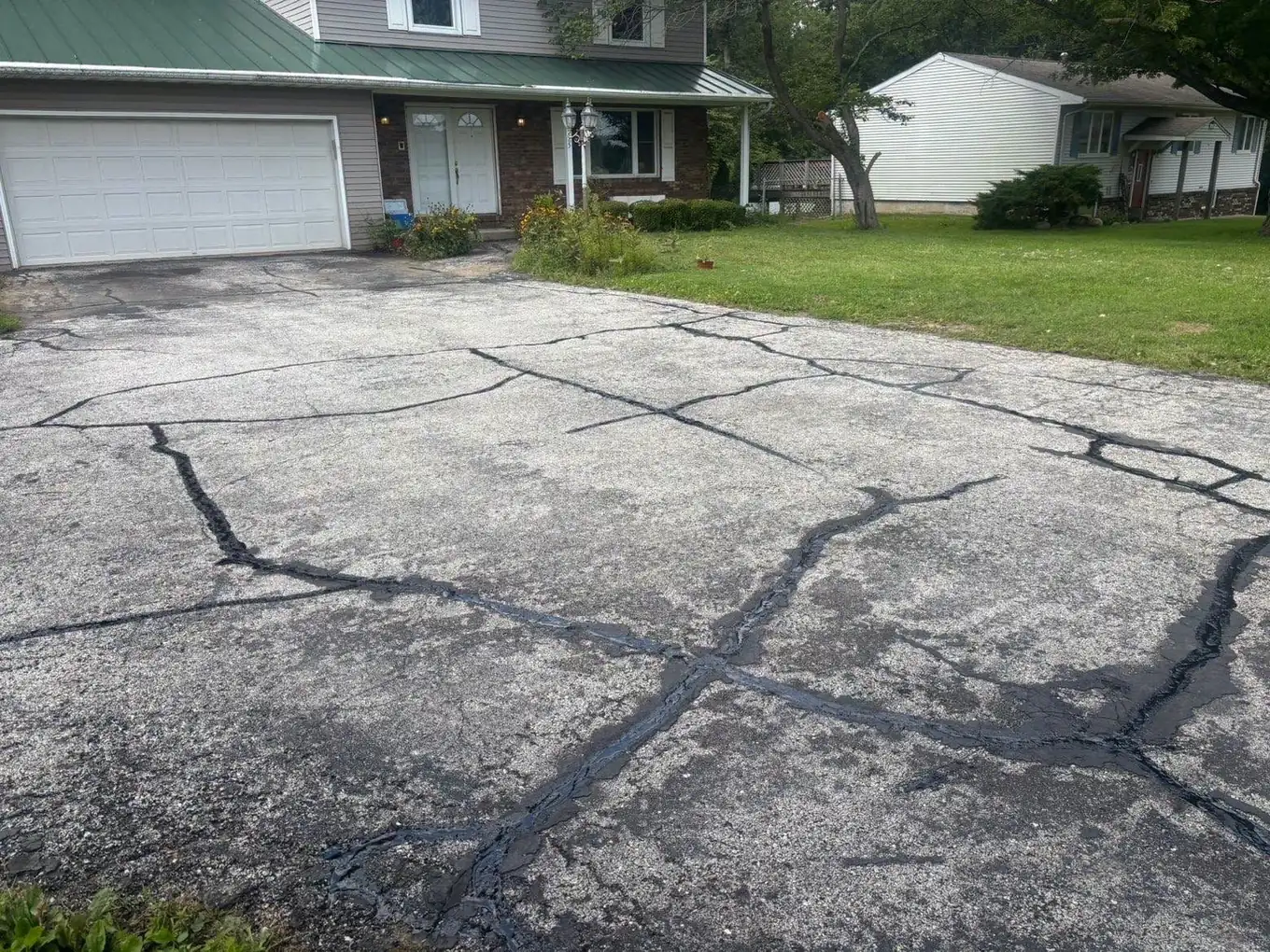 A wide residential asphalt driveway that is grey and oxidised with a network of open cracks.