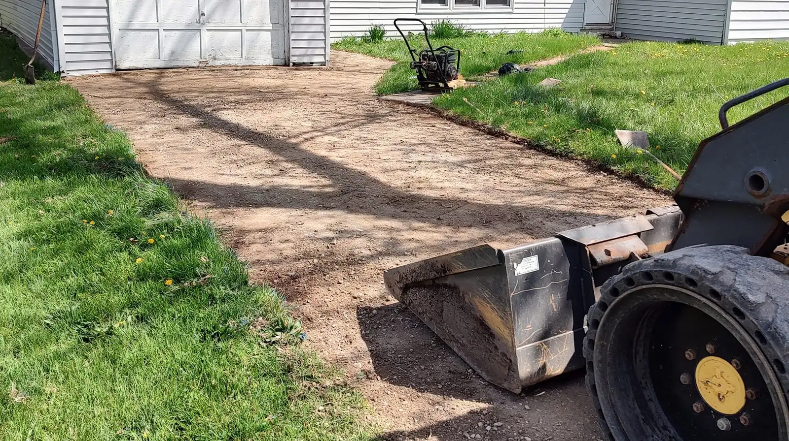 A skid steer bucket and a plate compactor sitting on a freshly graded and compacted driveway base beside a garage.