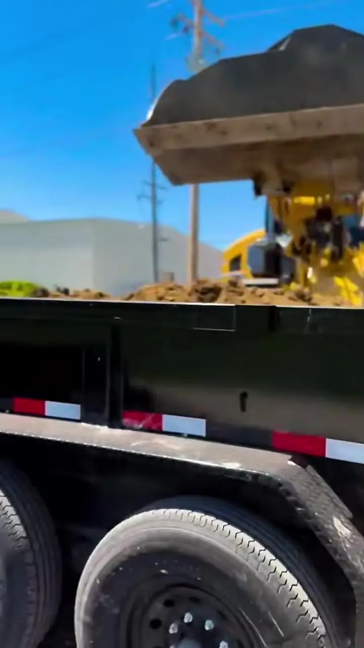 A wheel loader tipping a bucket of dark asphalt millings into a dump trailer under a clear sky.