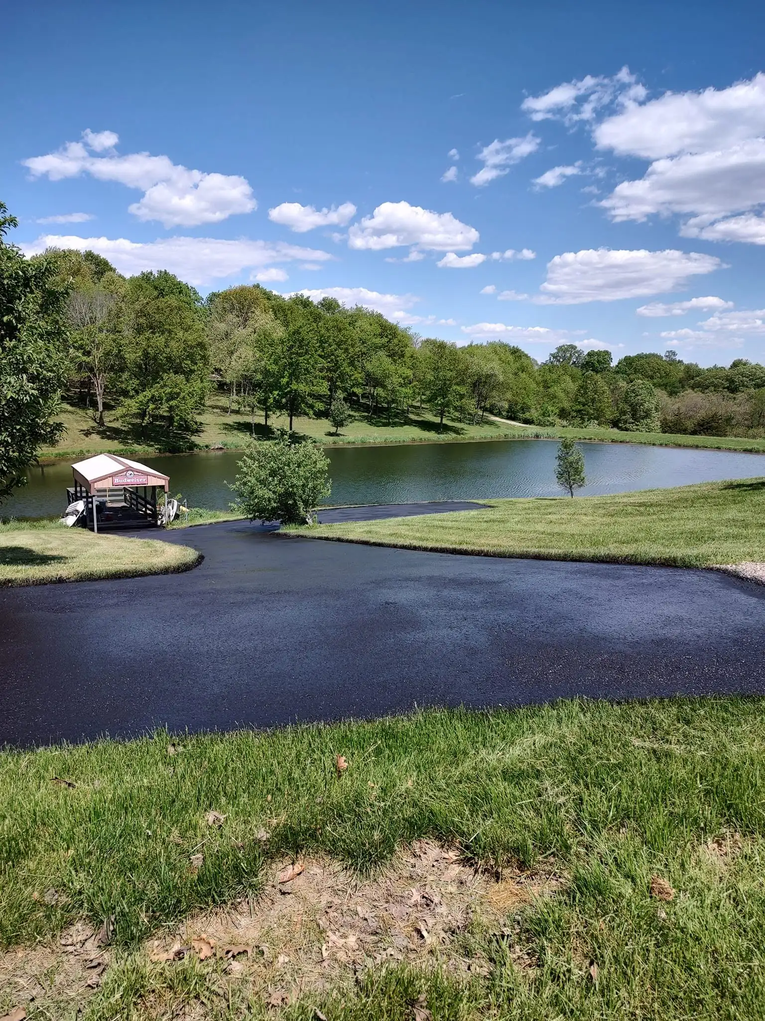 A deep black sealcoated drive curving around a private lake with a covered boat dock.