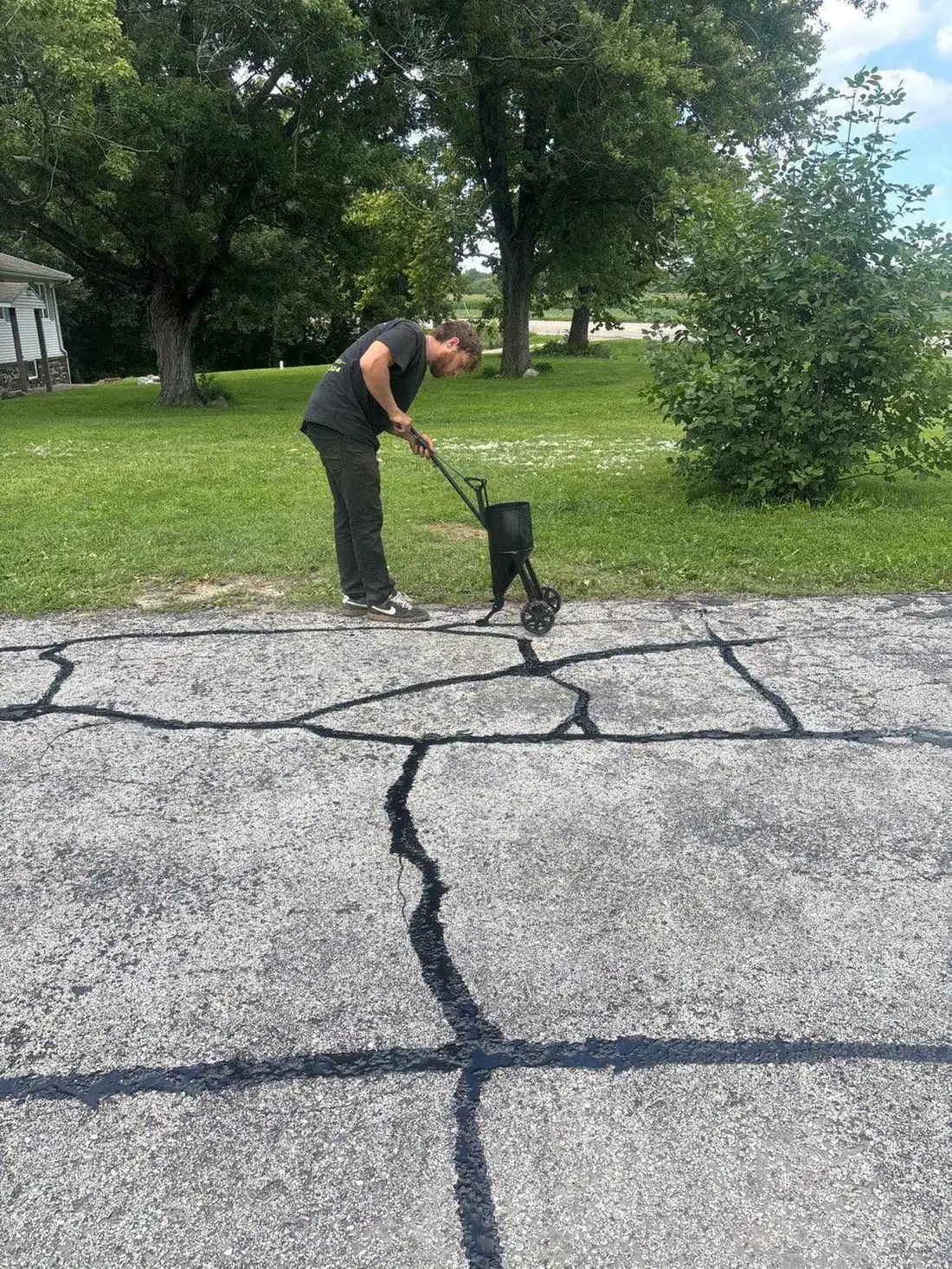 A J&N crew member walking a crack-filler pour pot along the seams of a cracked asphalt driveway.